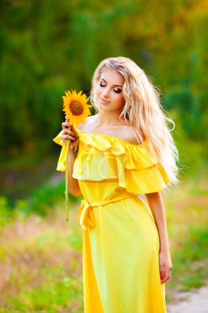 Foto de Hermosa joven rubia en vestido amarillo disfrutando de la naturaleza. Feliz hembra sonriente de pie en el bosque verde con girasoles. Una foto cálida. Obra artística. - Imagen libre de derechos