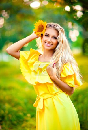 Foto de Hermosa joven rubia en vestido amarillo disfrutando de la naturaleza. Feliz hembra sonriente de pie en el bosque verde con girasoles. Una foto cálida. Obra artística. - Imagen libre de derechos