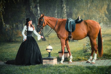Foto de Retrato de magnífica chica gótica de moda caminando un caballo. Fantasía obra de arte y hada. Increíble modelo morena en vestido blanco y negro y sombrero posando. - Imagen libre de derechos