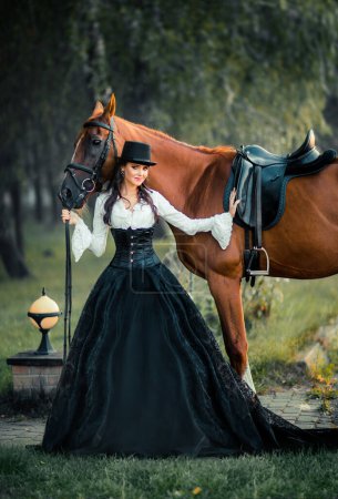 Foto de Retrato de magnífica chica gótica de moda caminando un caballo. Fantasía obra de arte y hada. Increíble modelo morena en vestido blanco y negro y sombrero posando. - Imagen libre de derechos