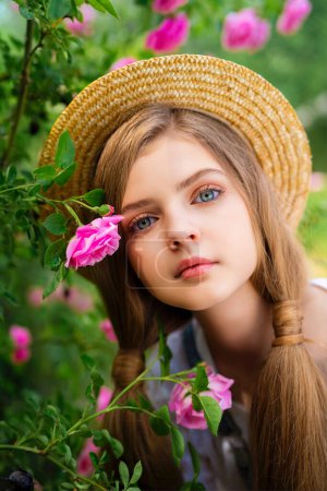 Schöne geflochtene blonde Mädchen in Jeans-Overalls und Strohhut genießen die Natur im Rosengarten. Junge hübsche Modell posiert in der Nähe Zaun mit grünen Bäumen und rosa Rosen auf background.Close up Porträt