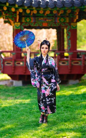 Foto de Hermosa chica morena asiática con palos en el pelo y kimono posando en el jardín de flores cerca de la pagoda. Retratos de primavera de la joven modelo de Japón. - Imagen libre de derechos