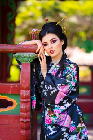 Téléchargez les photos : Belle fille brune asiatique avec des bâtons dans les cheveux et kimono posant dans le jardin fleuri près de la pagode. Portraits printaniers du jeune modèle japonais. - en image libre de droit