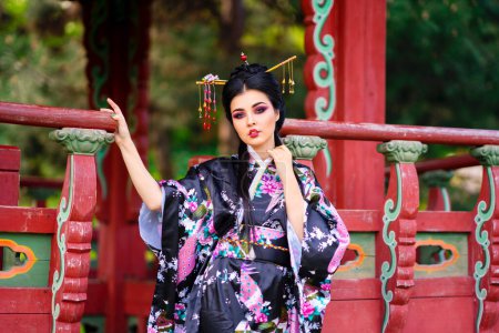 Photo for Beautiful asian brunette girl with sticks in hair and kimono posing in blossom garden near pagoda. Spring portraits of young Japan model. - Royalty Free Image