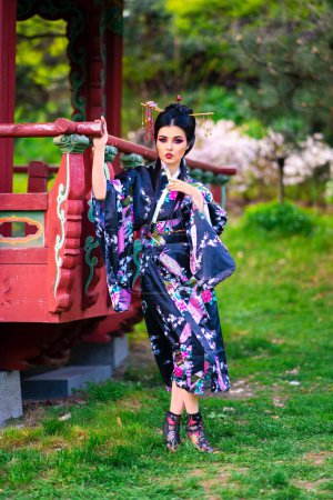 Téléchargez les photos : Belle fille brune asiatique avec des bâtons dans les cheveux et kimono posant dans le jardin fleuri près de la pagode. Portraits printaniers du jeune modèle japonais. - en image libre de droit