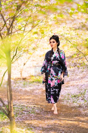Téléchargez les photos : Belle fille brune asiatique avec des bâtons dans les cheveux et kimono posant dans le jardin fleuri. Portraits printaniers du jeune modèle japonais. - en image libre de droit