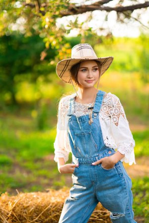Téléchargez les photos : Belle cow-girl blonde en chapeau de paille, chemise à carreaux et denim assis sur un fond vert nature. Oeuvre d'art colorée. - en image libre de droit