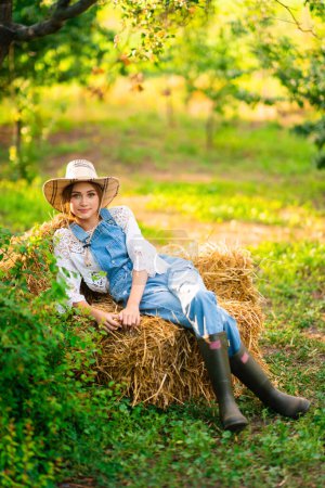 Foto de Hermosa vaquera rubia con sombrero de paja, camisa a cuadros y denim en general sentado sobre una paja en el fondo verde de la naturaleza. Colorida obra de arte. - Imagen libre de derechos