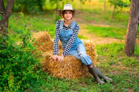 Photo for Beautiful blonde cowgirl in straw hat, checkered shirt and denim overall sitting on a straw on green nature background. Colorful art work. - Royalty Free Image