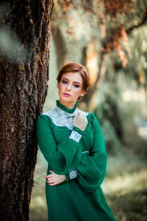 Beautiful red haired girl in green long vintage dress and basket of flowers standing near fence.Pretty tenderness model with perfect hairstyle dreaming and looking at camera.Art work and fairytale