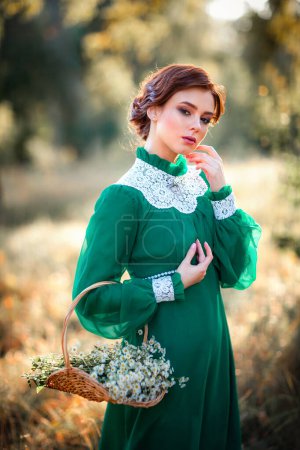 Beautiful red haired girl in green long vintage dress and basket of flowers standing near fence.Pretty tenderness model with perfect hairstyle dreaming and looking at camera.Art work and fairytale