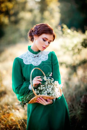 Schöne rothaarige Mädchen in grünen langen Vintage-Kleid und Korb mit Blumen in der Nähe von Zaun stehen.Hübsche Zärtlichkeit Modell mit perfekter Frisur träumen und in die Ferne schauen.Kunstwerk und Märchen