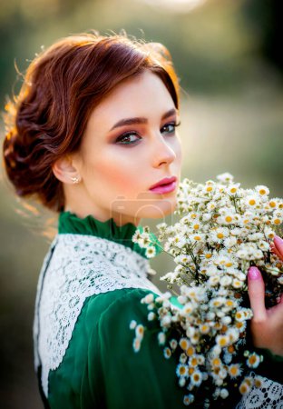Schöne rothaarige Mädchen in grünen langen Vintage-Kleid und Korb mit Blumen in der Nähe von Zaun stehen.Hübsche Zärtlichkeit Modell mit perfekter Frisur träumen und Blick auf camera.Art Arbeit und Märchen