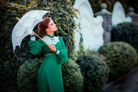 Schöne rothaarige Mädchen in grünen langen Vintage-Kleid und Korb mit Blumen stehen mit Spitzenschirmen. Hübsches Zärtlichkeit Modell mit perfekter Frisur träumen und in die Ferne schauen.Kunstwerk und Märchen