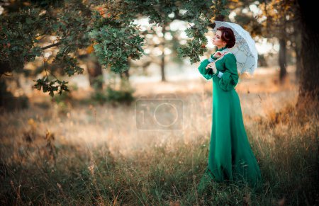Schöne rothaarige Mädchen in grünen langen Vintage-Kleid und Korb mit Blumen stehen mit Spitzenschirmen. Hübsches Zärtlichkeit Modell mit perfekter Frisur träumen und in die Ferne schauen.Kunstwerk und Märchen