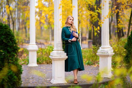 Téléchargez les photos : Belle jeune fille blonde bouclée en vert longue robe vintage et dentelle debout près de l'alcôve dans le parc d'automne.Jolie modèle de tendresse posant et regardant loin .Oeuvre d'art et conte de fées - en image libre de droit