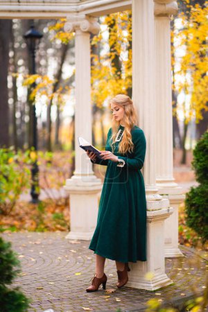 Téléchargez les photos : Belle jeune fille blonde bouclée en vert longue robe vintage et dentelle debout près de l'alcôve dans le parc d'automne et le livre de lecture.Jolie modèle de tendresse posant. Oeuvre d'art et conte de fées - en image libre de droit