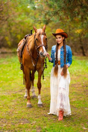Photo for Beautiful cowgirl with extra long braided hair in vintage lace dress and orange hat posing on ranch with horse. Portrait of a pretty model in countryside. Warm art. - Royalty Free Image