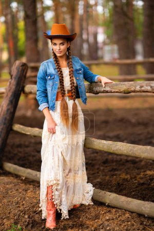 Photo for Beautiful cowgirl with extra long braided hair in vintage lace dress and orange hat posing on ranch. Portrait of a pretty model in countryside. Warm art. - Royalty Free Image