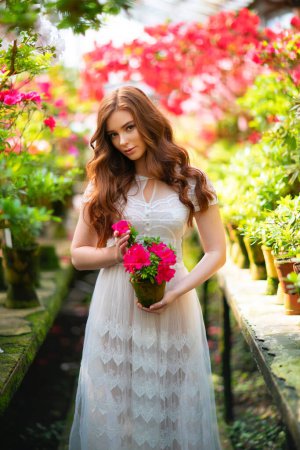 Belle fille aux cheveux rouges dans une robe en dentelle blanche debout dans un jardin avec des fleurs colorées. Oeuvre d'art de femme romantique. Jolie modèle de tendresse regardant la caméra.