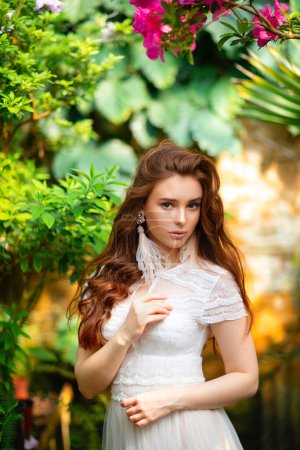 Téléchargez les photos : Belle fille aux cheveux rouges dans une robe en dentelle blanche debout dans un jardin avec des fleurs colorées. Oeuvre d'art de femme romantique. Jolie modèle de tendresse regardant la caméra. - en image libre de droit