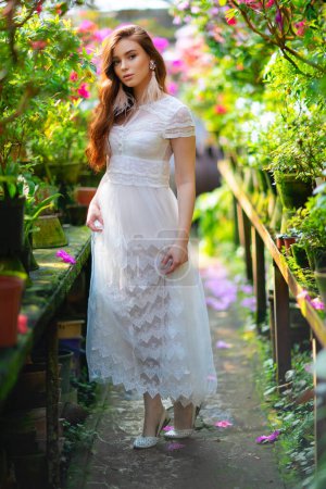 Téléchargez les photos : Belle fille aux cheveux rouges dans une robe en dentelle blanche debout dans un jardin avec des fleurs colorées. Oeuvre d'art de femme romantique. Jolie modèle de tendresse regardant la caméra. - en image libre de droit