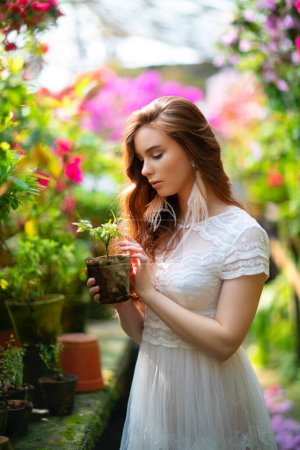 Photo for Beautiful red haired girl in a white lace dress standing in a garden with colorful flowers. Art work of romantic woman .Pretty tenderness model looking at flower. - Royalty Free Image