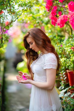 Belle fille aux cheveux rouges dans une robe en dentelle blanche debout dans un jardin avec des fleurs colorées. Oeuvre d'art de femme romantique. Jolie modèle de tendresse regardant la fleur.