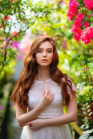 Schöne rothaarige Mädchen in einem weißen Spitzenkleid steht in einem Garten mit bunten Blumen. Kunstwerk der romantischen Frau. Schöne Zärtlichkeit Modell Blick in die Kamera.