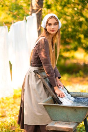 Bastante joven lavandera en traje medieval lavado de vestidos en antiguo abrevadero en la naturaleza en un pueblo.Hermosa chica que trabaja en el campo. Obra de arte de cuento.
