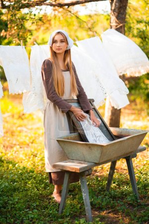 Téléchargez les photos : Jolie jeune blanchisseuse en costume médiéval lavant des robes dans une vieille cuvette sur la nature dans un village.Belle fille travaillant à la campagne. Œuvre d'art conte de fées. - en image libre de droit
