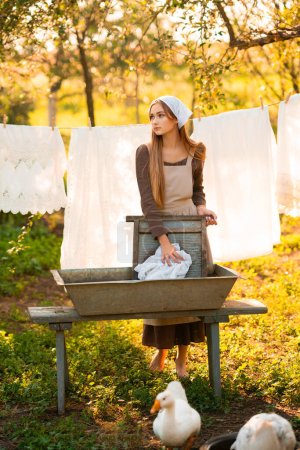 Photo for Pretty young laundress in medieval costume washing dresses in old trough on nature in a village.Beautiful girl working in countryside. Fairytale art work. - Royalty Free Image