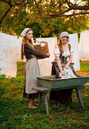Téléchargez les photos : Deux jolies jeunes blanchisseries en costume médiéval lavant des robes dans une vieille cuvette sur la nature dans un village.Belles filles qui travaillent à la campagne. Œuvre d'art conte de fées. - en image libre de droit