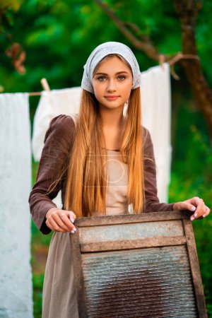 Pretty young laundress in medieval costume washing dresses in old trough on nature in a village.Beautiful girl working in countryside. Fairytale art work.