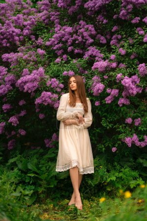 Foto de Hermosa chica con el pelo largo y vestido blanco vintage posando en el jardín lila. Modelo romántico disfrutando de la naturaleza. - Imagen libre de derechos