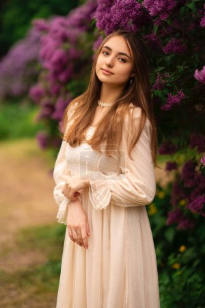 Téléchargez les photos : Belle fille aux cheveux longs et robe blanche vintage posant dans un jardin lilas. Modèle romantique jouissant de la nature. - en image libre de droit