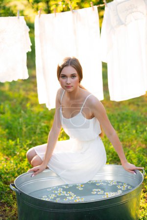Photo for Beautiful blonde long hair girl in white dress sitting in a trough with water and chamomile. Warm art work. Young model take a bath on nature with vintage clothes as background. - Royalty Free Image