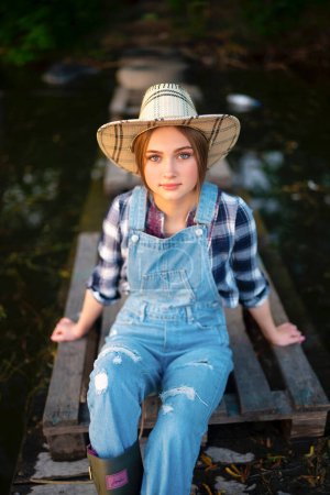 Photo for Portrait of beautiful blonde girl in straw hat, denim overall, checkered shirt and rubber boots fishing. Young model sitting on wood berth near river  with fishing rod. Colorful art work on nature. - Royalty Free Image