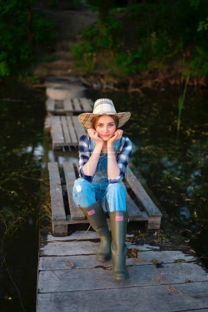 Foto de Retrato de hermosa chica rubia en sombrero de paja, mezclilla en general, camisa a cuadros y botas de goma de pesca. Modelo joven sentado en el amarradero de madera cerca del río con caña de pescar. Colorida obra de arte sobre la naturaleza. - Imagen libre de derechos