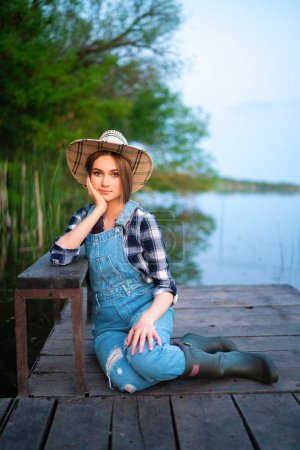 Photo for Portrait of beautiful blonde girl in straw hat, denim overall, checkered shirt and rubber boots fishing. Young model sitting on wood berth near river  with fishing rod. Colorful art work on nature. - Royalty Free Image