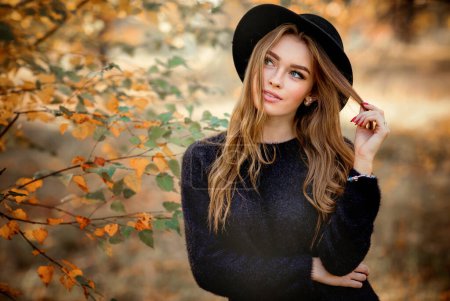 Téléchargez les photos : Gros plan portrait d'une belle fille en robe sombre et chapeau noir debout près de feuilles d'automne colorées. Oeuvre d'art de femme romantique. Jolie modèle de tendresse détournant les yeux. - en image libre de droit