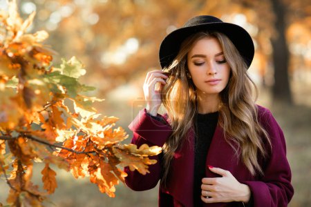 Téléchargez les photos : Gros plan portrait d'une belle fille en robe sombre et chapeau noir debout près de feuilles d'automne colorées. Oeuvre d'art de femme romantique. Jolie modèle de tendresse regardant vers le bas. - en image libre de droit