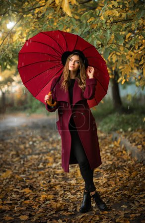 Téléchargez les photos : Gros plan portrait d'une belle fille en robe bordeaux et chapeau noir debout près de feuilles d'automne colorées avec parapluie. Oeuvre d'art de femme romantique. Jolie modèle de tendresse détournant les yeux. - en image libre de droit