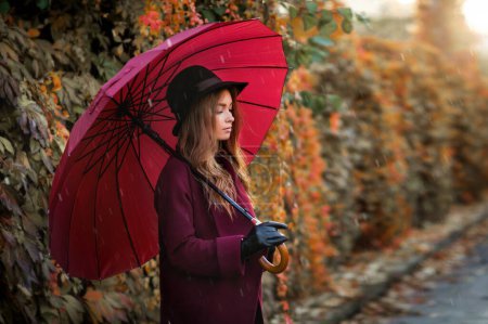 Foto de Retrato de una hermosa chica en vestido de borgoña y sombrero negro de pie cerca de hojas de otoño de colores con paraguas. Obra de arte de mujer romántica . - Imagen libre de derechos