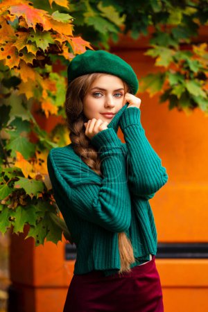 Téléchargez les photos : Portrait de jeune belle fille blonde aux yeux bleus et aux cheveux tressés.Modèle attrayant en pull tricoté vert et béret debout près de vieux bus orange rétro avec fond d'automne. Art coloré. - en image libre de droit