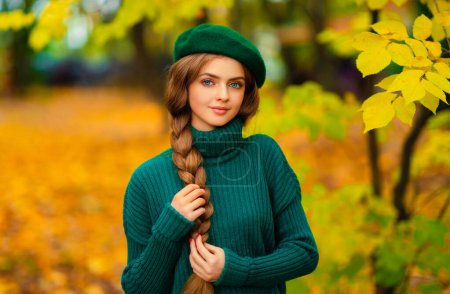 Foto de Retrato de joven hermosa rubia con ojos azules y cabello trenzado.Modelo atractivo en suéter de punto verde y boina de pie en la naturaleza de otoño. Arte colorido. - Imagen libre de derechos