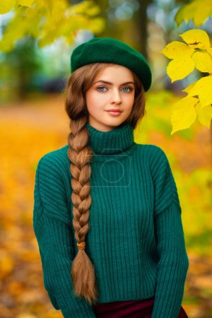 Téléchargez les photos : Portrait de jeune belle fille blonde aux yeux bleus et aux cheveux tressés.Modèle attrayant en pull tricoté vert et béret debout dans la nature automnale. Art coloré. - en image libre de droit