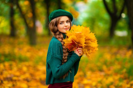 Photo for Portrait of young beautiful blonde girl with blue eyes and braided hair.Attractive model in green knitted sweater and beret standing in autumn nature and holding bouquet with leaves. Colorfull art. - Royalty Free Image