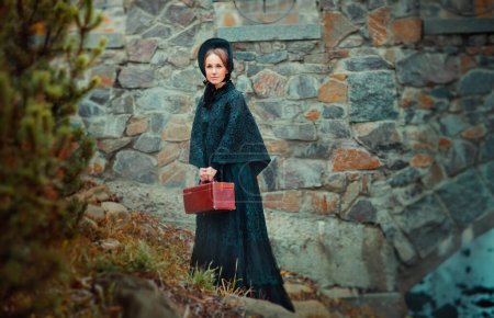 Beautiful woman in black long vintage dress and hood standing near stone wall. Art work of romantic woman with a valise. Pretty tenderness model dreaming and looking afar. Woman traveling.