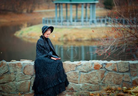 Beautiful woman in black long vintage dress and hood sitting on stone wall. Art work of romantic woman with a valise. Pretty tenderness model dreaming and looking down. Woman with a book.